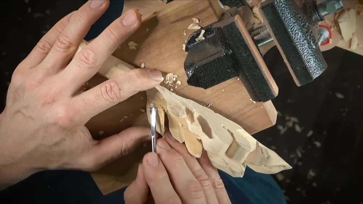 Both hands guide a short tool along a thin carved leaf next to the vise