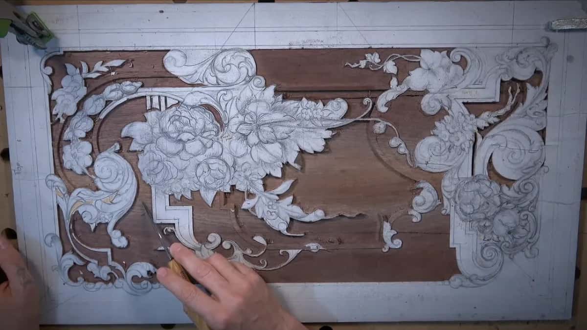 Top view of the walnut panel with its background taken down, paper still on the scrolls and flowers, a tool in hand